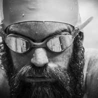 Black and white headshot of man with a beard wearing swim goggles and hat covered in water droplets.
