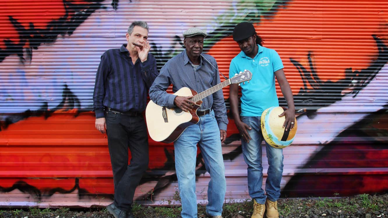 3 men with a guitar and mouthorgan are leaning against a wall painted with red and purple graffiti.