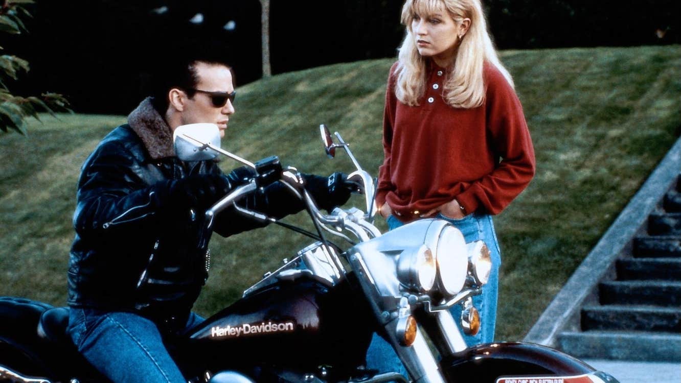 A young woman is speaking to a young man on a motorcycle.