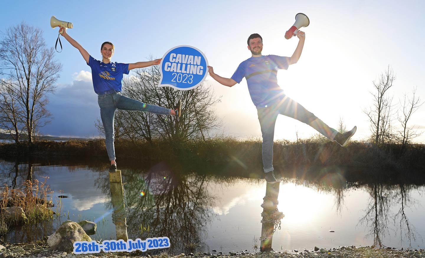 A man and a woman standing on a post in some water with the other leg and arms in the air holding between them a sign in blue and white with the event title on, set outside in the countryside with the sun shining behind them.