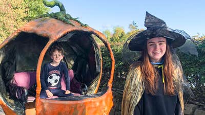 A smiling girl in black witches hat and costume with orange pumpkin style small carriage behind with young boy seated in it.