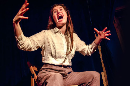 A woman with long hair is seated on a chair with arms and hands spread out, appearing to be screaming or shouting, against black background.