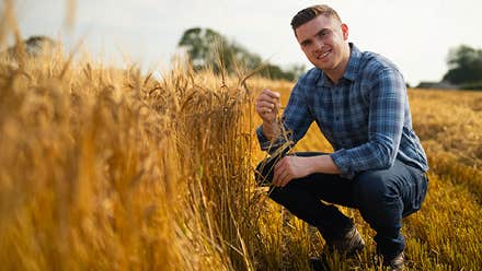 A man crouched down in a field beside golden wheat.