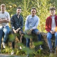 6 casually dressed men seated on a low stone wall surrounded by greenery