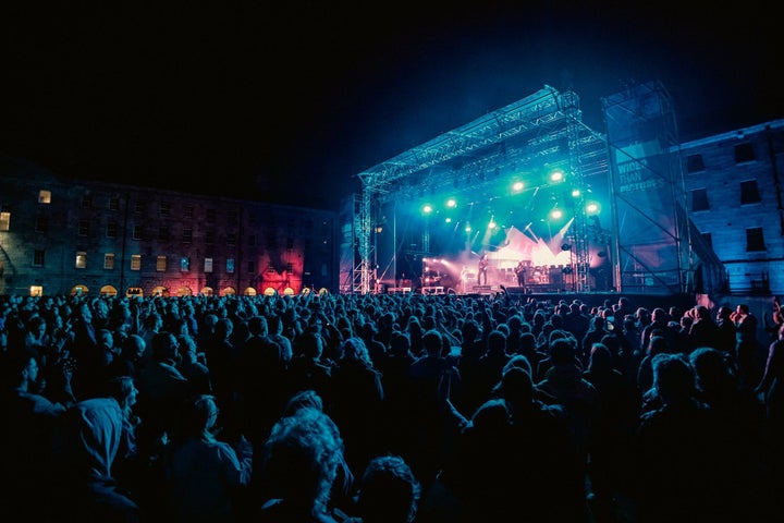 Distant view of large outdoor stage at night with large audience watching musicians