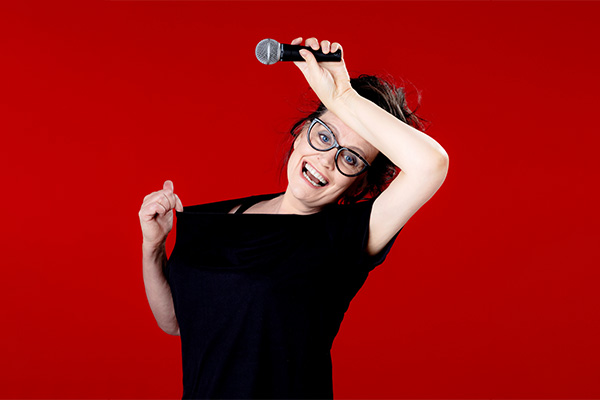 ANNE GILDEA: HOW TO GET THE MENOPAUSE AND ENJOY IT. A woman with a wide smile holding the back of her left hand up to her forehead with a mic in it and her right hand pulling away the neck of a black top as if too hot, against a red background.