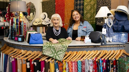 Two people behind a shop counter with scarfs on hangers hanging from a rail on it