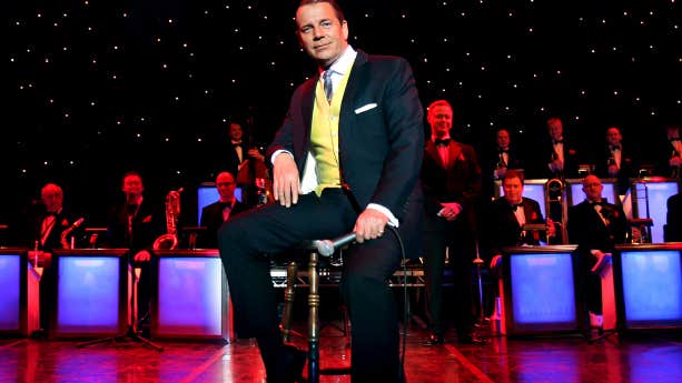 Sinatra's Greatest Hits with Stephen Triffitt. A man in black suit and yellow waistcoat is seated on a stool holding a mic on a stage with band behind him.