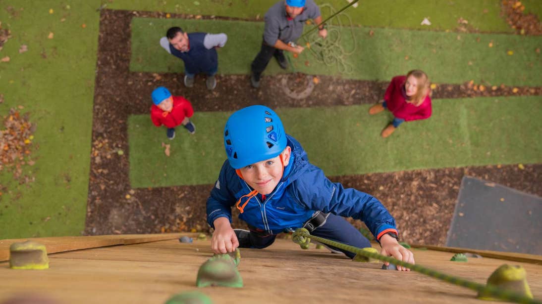 Young boy smiling at the camera while on a wooden climbing wall with people watching from below.