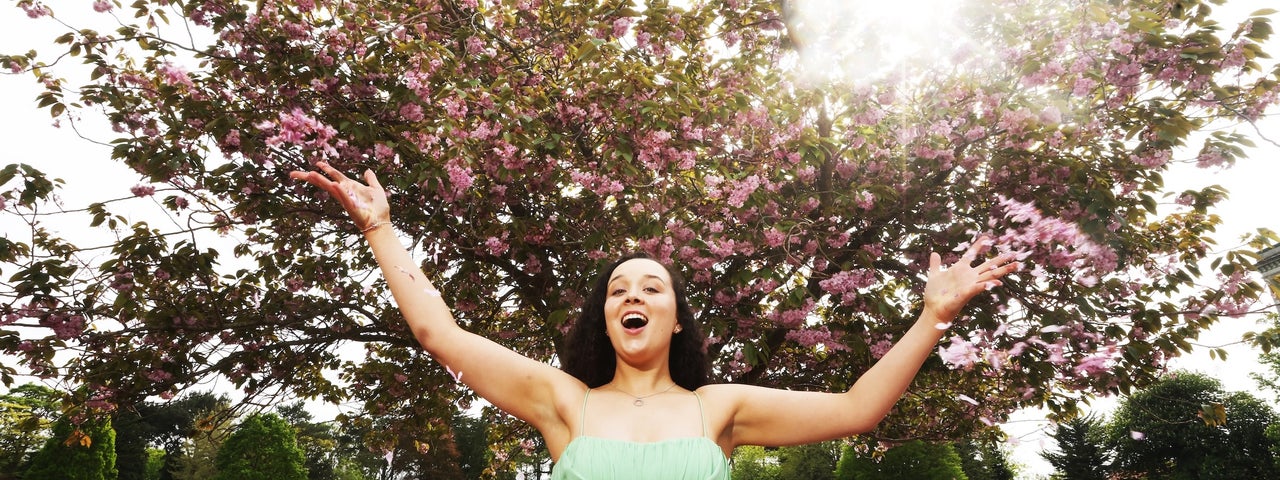 A woman in a green dress with arms outstretched, singing with trees in the background in blossom.