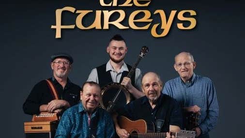 Group photo of The Fureys, 3 standing, 2 seated in front, holding musical instruments, smiling at camera against dark background with band name above in dull yellow/gold text.