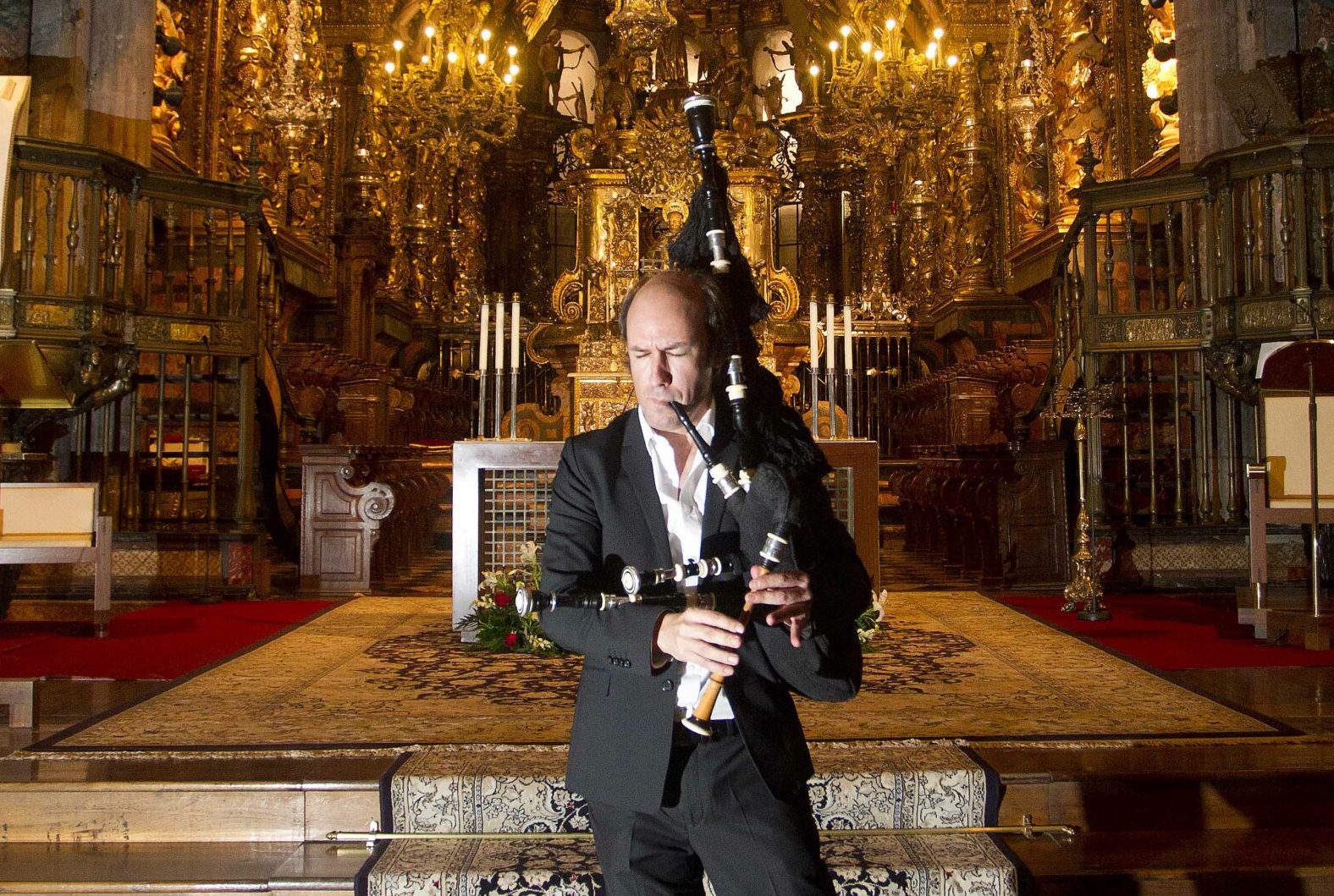 A man in shirt and jacket is playing pipes standing in front of ornate, gold alter and surroundings in a church.