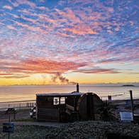A coastal sauna overlooking a calm sunrise sky filled with pink and orange clouds