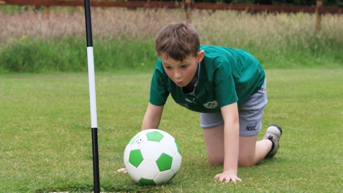 A child on their hands and knees blowing on a football to get it in a hole