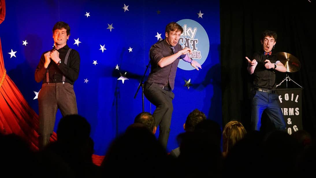 Three men performing on stage in front of a packed audience at the Cat Laughs Festival
