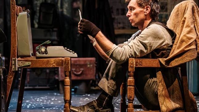 A man seated in a chair frame looking at a piece of paper with old small typewriter on an old chair in front of him.
