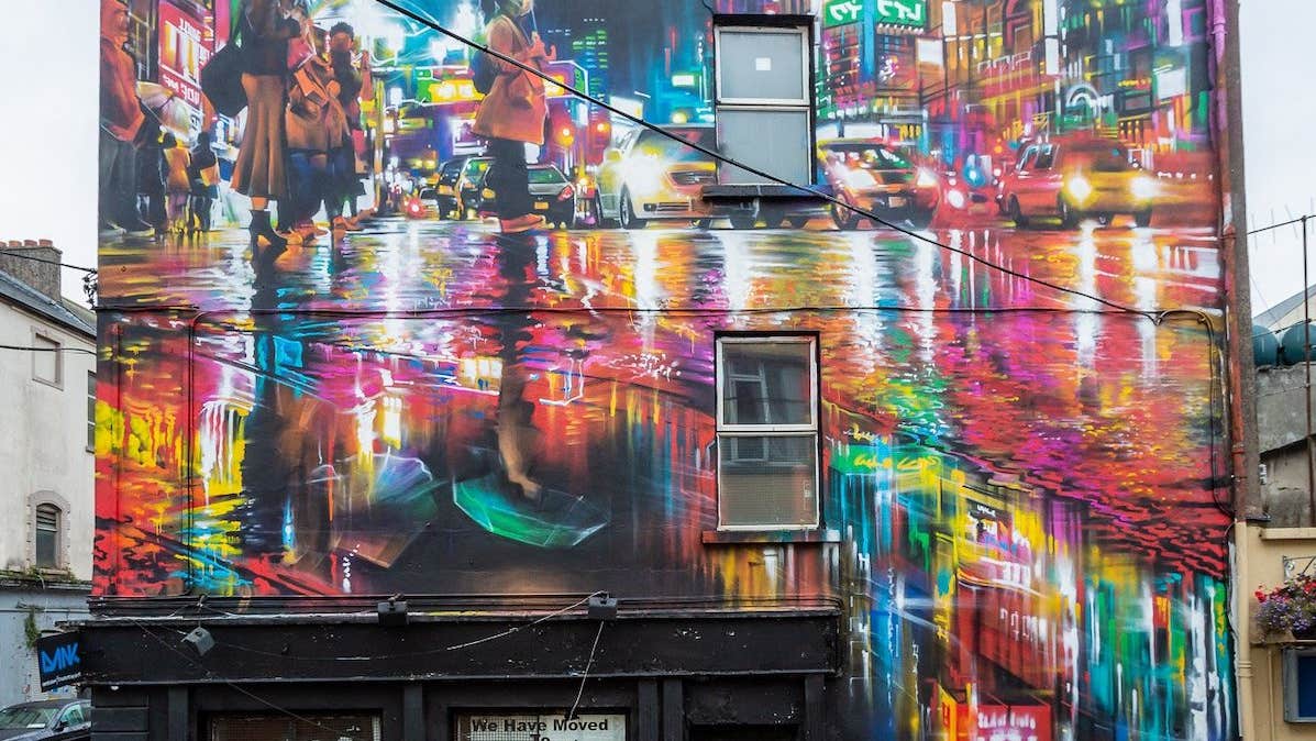 Waterford Walls 2018 - Dan Kitchner. Photographer - Karl Kachmarksy, side of a building painted with a brightly lit city scene at night with reflections in wet road.