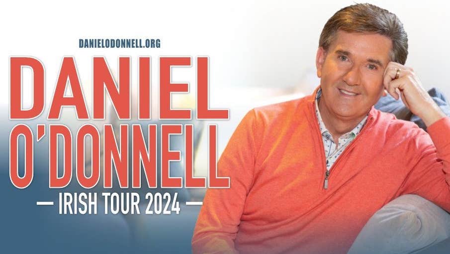 Daniel O'Donnell in concert. A gently smiling man in an orange jumper is resting his left arm on the arm of a sofa.