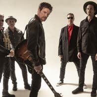 5 men dressed in dark clothes and leather jackets are standing in a casual group looking serious with one in the centre towards the front with a guitar swinging down looking questioningly