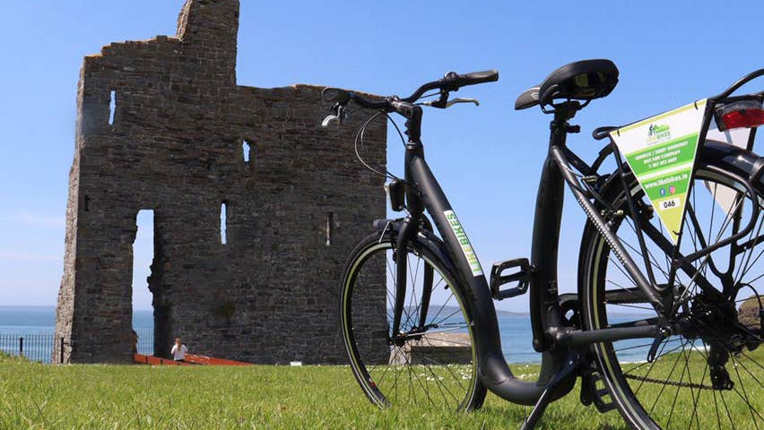 A lone bicycle propped up with the ruins of a small castle a short distance away with the coast in the background