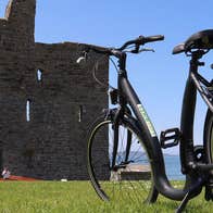 A lone bicycle propped up with the ruins of a small castle a short distance away with the coast in the background