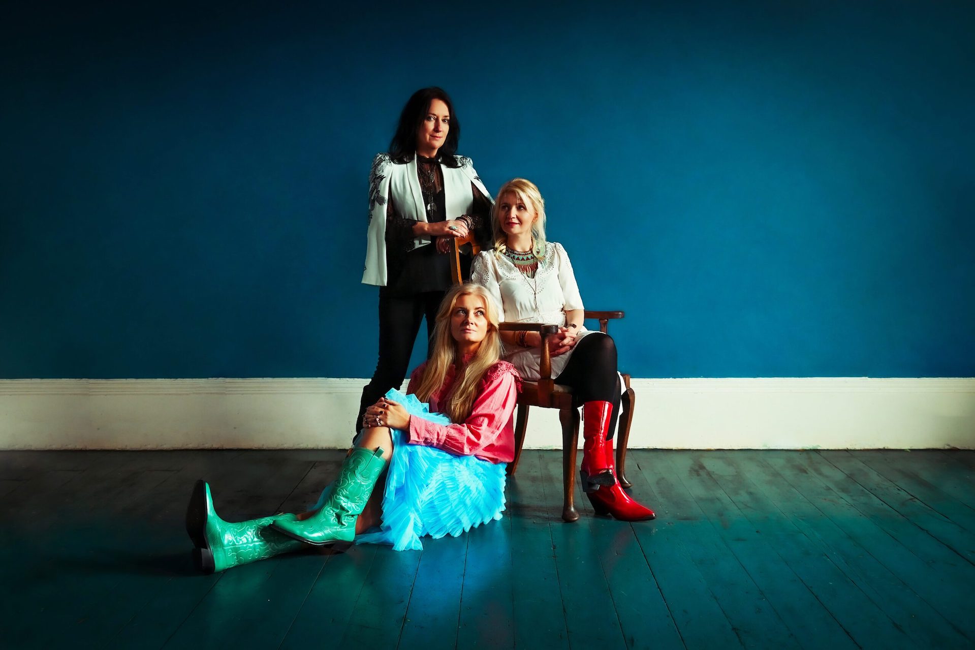 3 women, 1 standing, 1 seated in a wooden chair, 1 sat in front on a wooden floor, inside against dark blue wall.