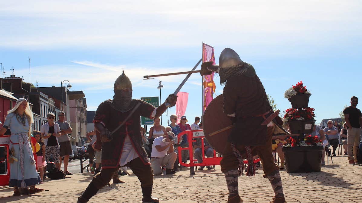 2 men dressed in viking type clothing in a mock battle with swords on a paved area with people watching in the background and blue sky.