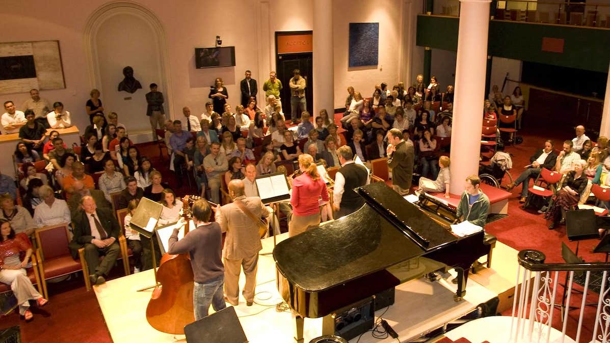 Viewed from behind and above, rear view of small stage with musicians playing and seated audience in a hallway with stairs