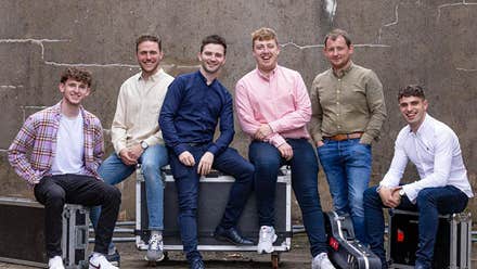 Line of 6 smiling men in different coloured shirts, seated casually on equipment boxes.