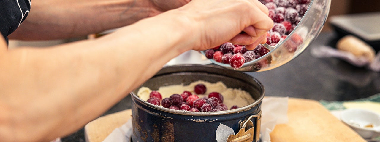 View of pair of hands holding a bowl of red berries, tipping them into a round, deep cake tin