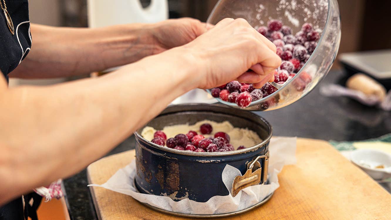 View of pair of hands holding a bowl of red berries, tipping them into a round, deep cake tin