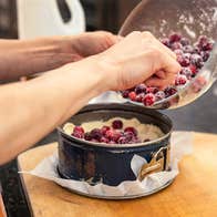 View of pair of hands holding a bowl of red berries, tipping them into a round, deep cake tin