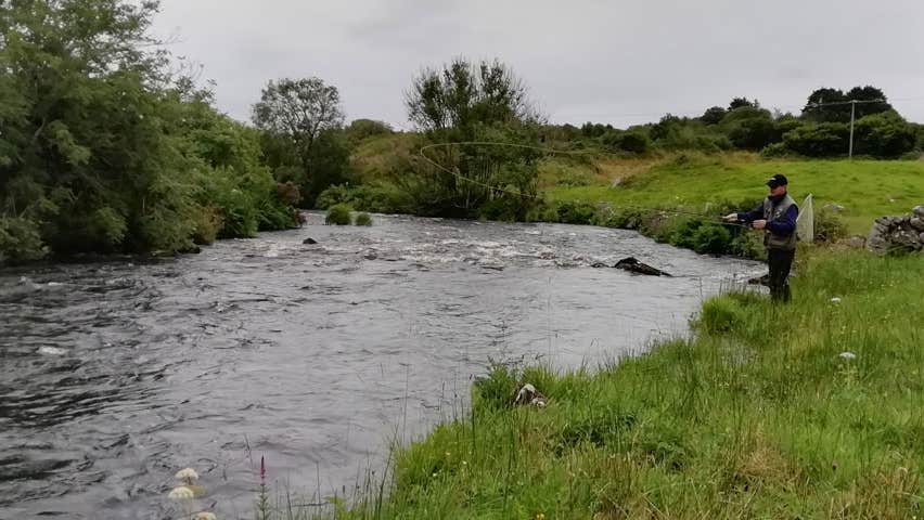 A lone fisherman standing on a river bank with fishing rod in hand
