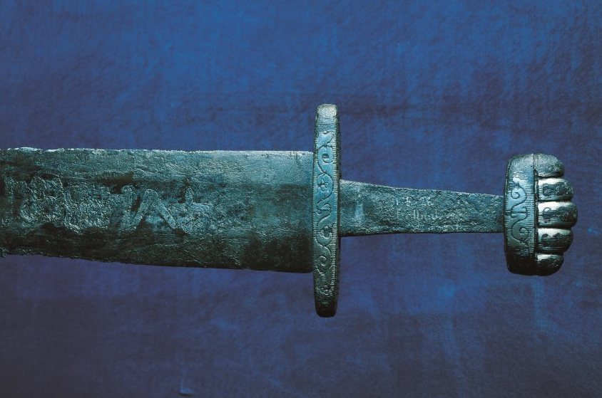 A Viking sword, aged metal green with gold detail on handle against dark blue background.