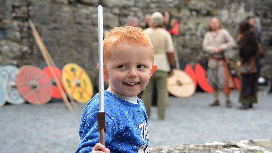 A young boy in a blue top holding up a small wooden sword, smiling looking away to the right, blurred background with adults in costume and shields leaning up against a stone wall.