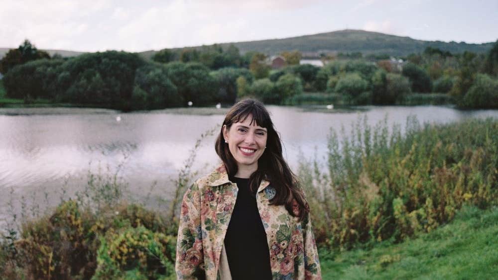 Photo Mitch Conlon. A smiling woman outdoors with a lake and bushes behind her.