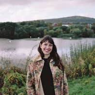 Photo Mitch Conlon. A smiling woman outdoors with a lake and bushes behind her.
