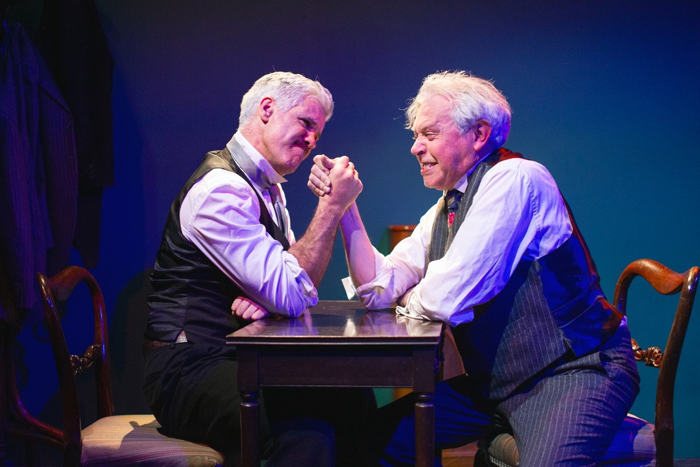 Counterparts & A Little Cloud. 2 older men in shirts and waistcoats are seated either side of a small wooden table engaged in an arm wrestling match.