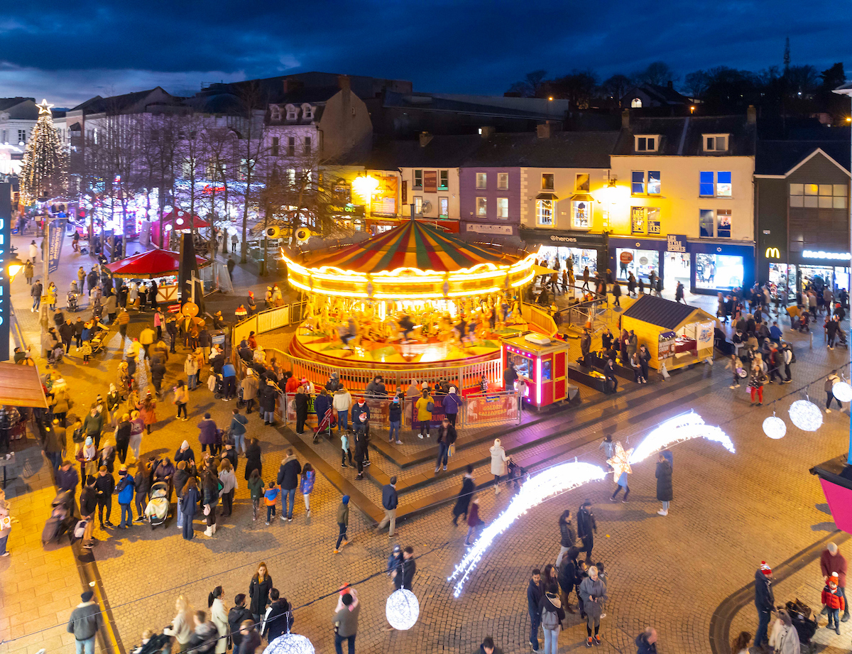 A large, moving, golden lit carousel is lit up at night in a town square with lots of people at night.