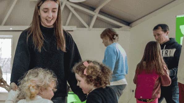 pop-up Gaeltacht at Ardgillan Castle - a smiling woman looking down on 2 young children all dancing in a circle with others in the background.