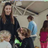 pop-up Gaeltacht at Ardgillan Castle - a smiling woman looking down on 2 young children all dancing in a circle with others in the background.
