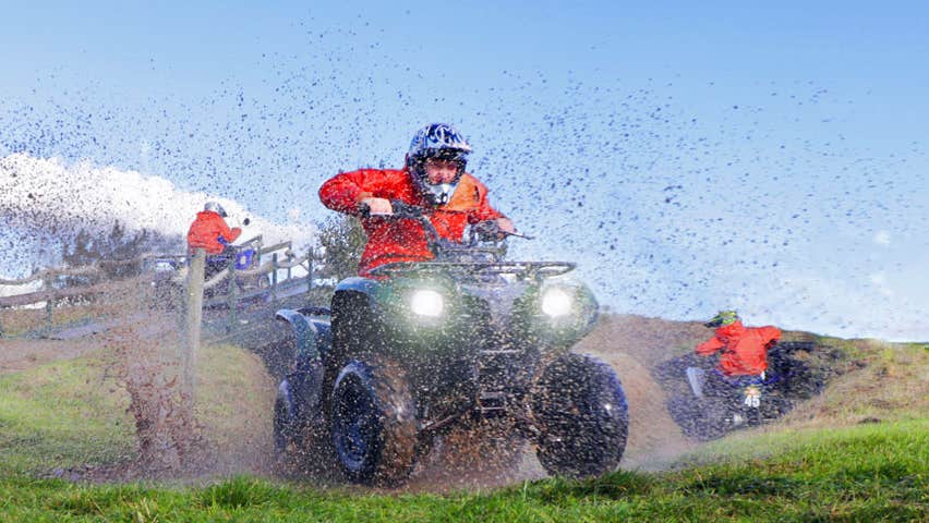 A man on a quad bike driving through a muddy field with mud spray visible in the image