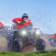 A man on a quad bike driving through a muddy field with mud spray visible in the image