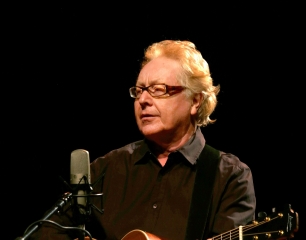 A man wearing a dark shirt holding a guitar with a large mic on a stand in front of him, against a black background.