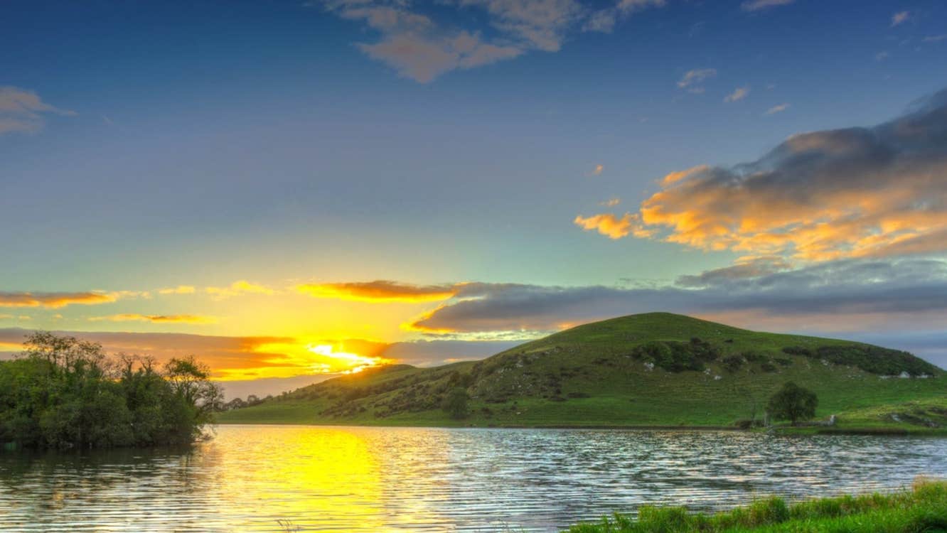Sun setting behind green hills beside a lake