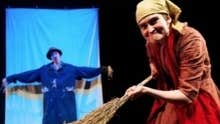 A woman acting as an old woman sweeping with a large twig broom.