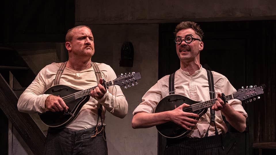 DRUID - Three Short Comedies at Watergate Theatre. 2 men in old style collarless shirts are playing banjos with animated faces on a stage.