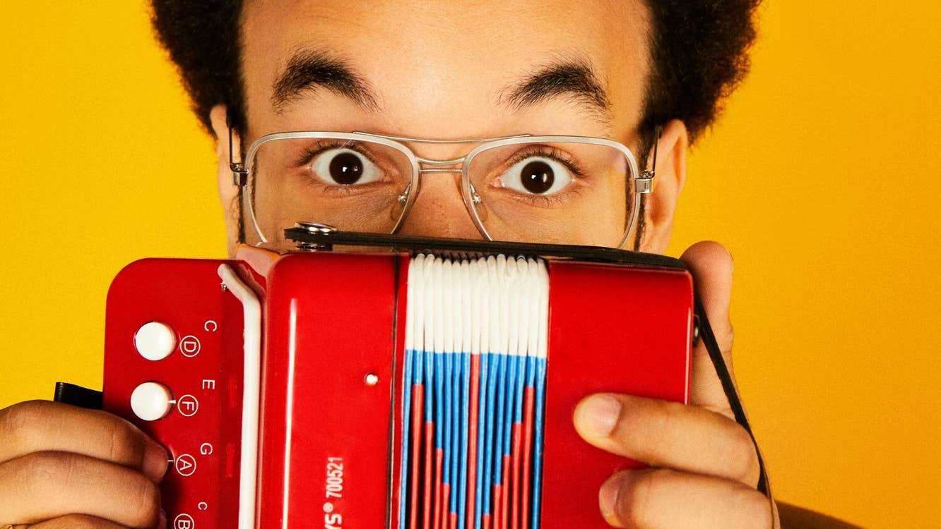 A man in glasses with raised eyebrows peering over the top of a small accordion he's holding up in front of him.