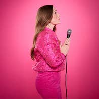 A woman dressed in pink is standing, viewed from the side with hands held in front of her in prayer holding a mic, against bright pink background.