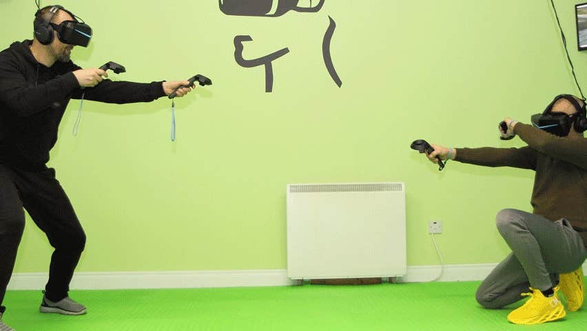 One man standing with vr equipment and one man crouching with vr equipment in a games room